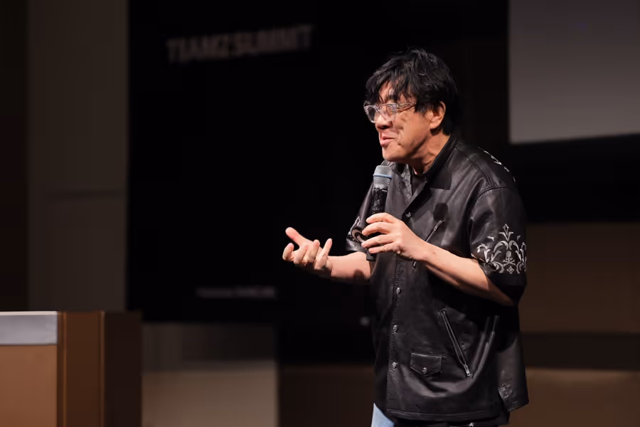 マイクを持って講演する黒いレザージャケットとメガネを着用した男性。