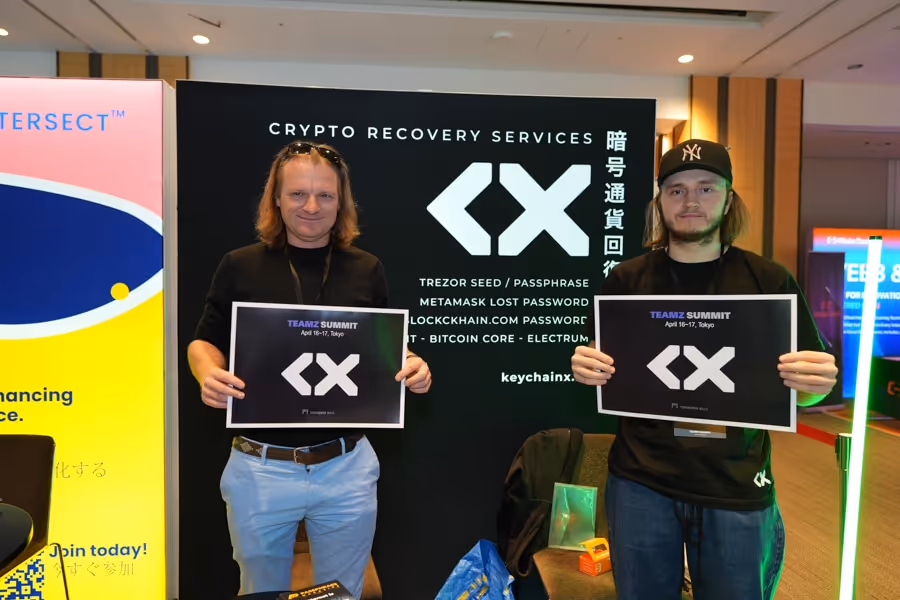 二人の男性が暗号通貨回復サービスの広告の前でTEAMZ SUMMITのポスターを持って立っている。