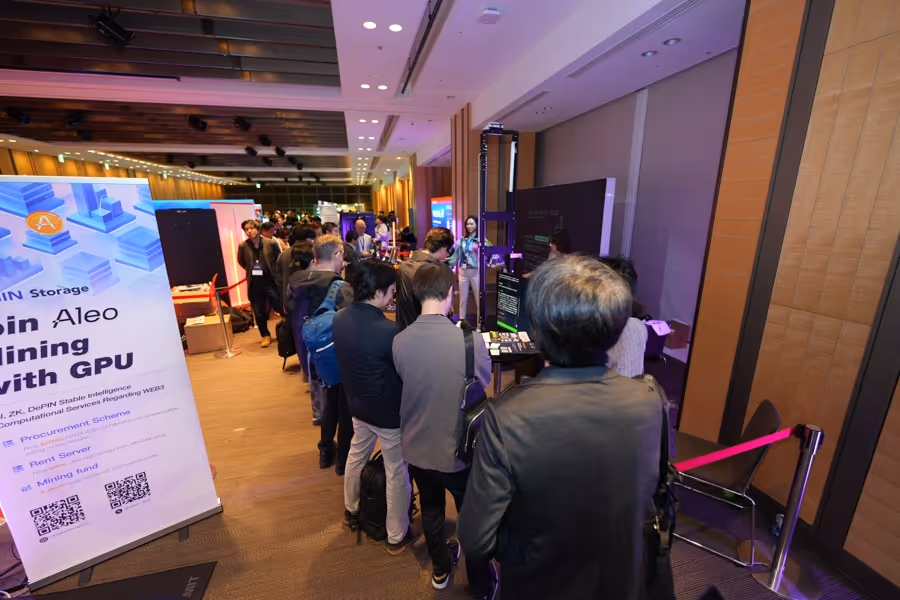 会場の展示ブースで、複数の人々がコンピューター画面や資料を見ながら順に並んでいる様子。