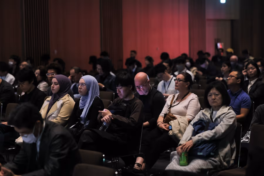 薄暗い会場で講演を真剣に聴く多様な聴衆の姿。