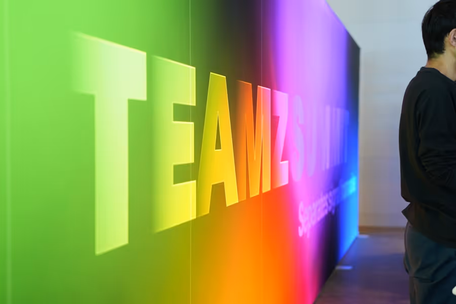 虹色の背景に大きく表示されたTEAMZSUNITEの文字と右側に立つ男性の一部。