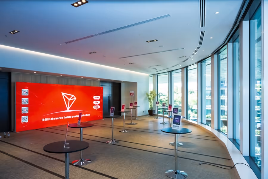 ガラス窓に沿って置かれた高いテーブルが並ぶ明るい会場の内観、壁の赤いディスプレイにはTRONのロゴとメッセージが表示されている。