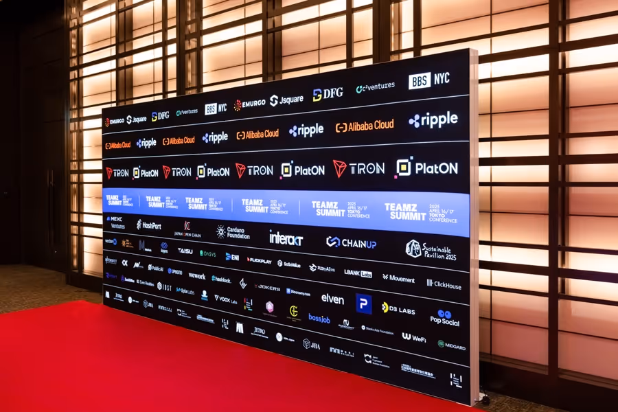 赤いカーペットの上に設置された、多数の企業ロゴが並ぶ黒い広告ボードが和風の照明壁の前にある。