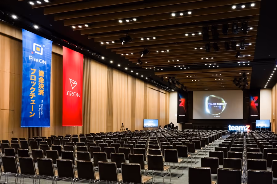 空の会議室、整然と並んだ椅子と前方の大きなスクリーンに『TEAMZ SUMMIT』の表示。