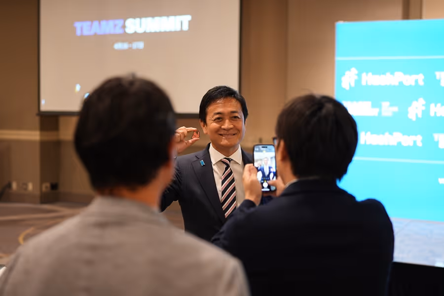 スーツ姿の男性がカメラに向かって微笑みながら指で小さな物を持ち、背後の人がその様子をスマートフォンで撮影している。