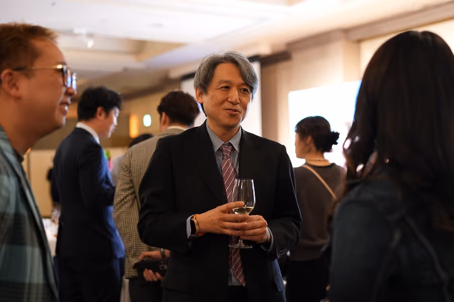 スーツを着た中年の男性がグラスを持ちながら他の人々と会話している屋内の社交イベント。