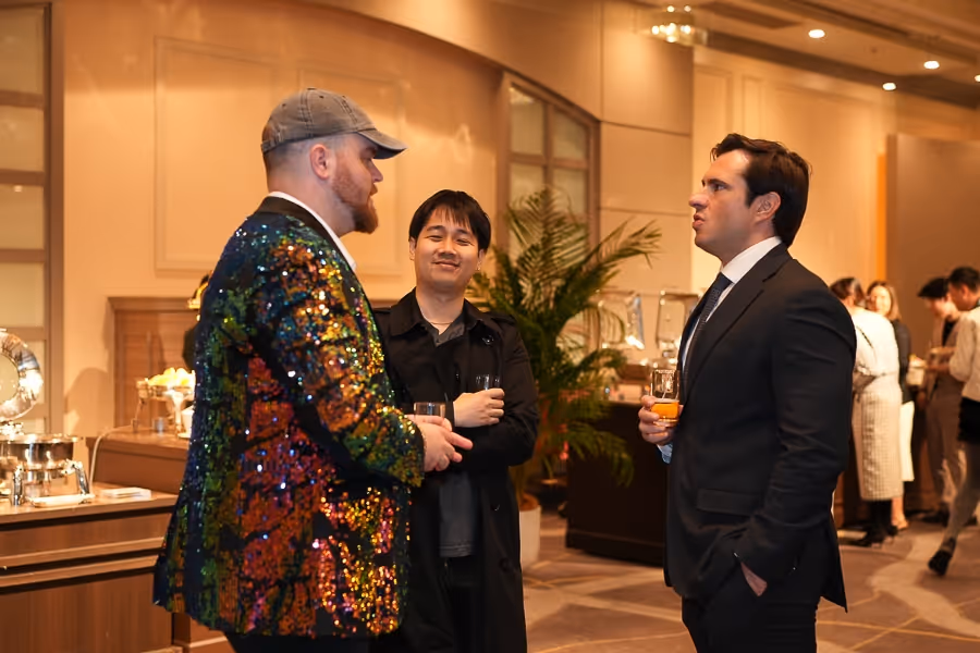 カクテルグラスを持ち、華やかなスパンコールジャケットの男性、黒のコートを着た男性、スーツ姿の男性が会話している室内のイベントシーン。