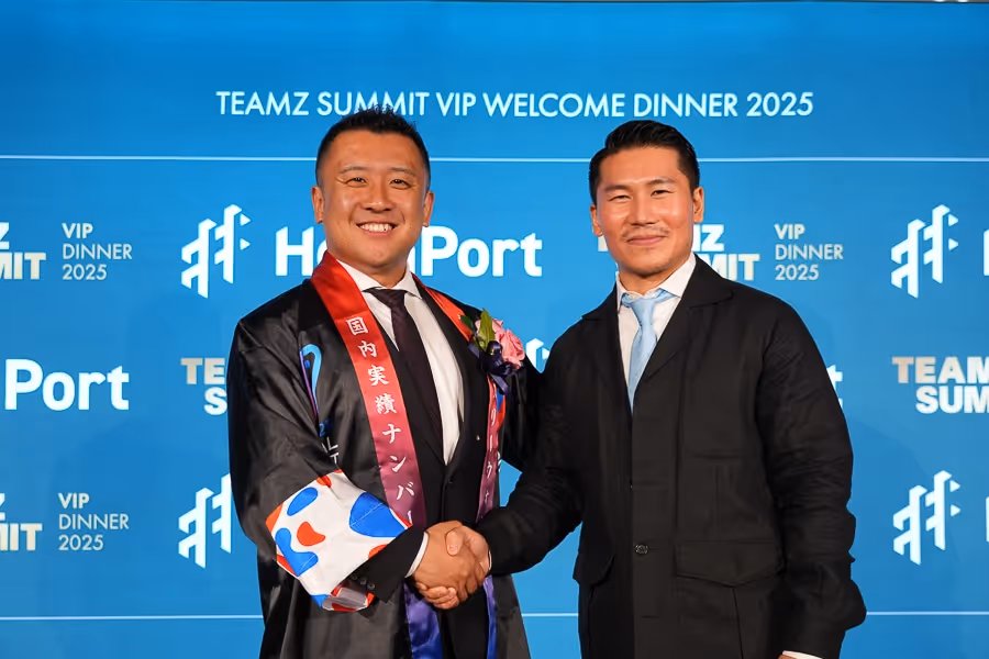 スーツに花をつけた男性と黒いジャケットの男性が、青い背景の前で握手をして笑顔を見せている。