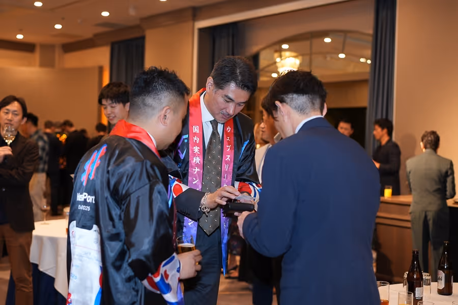 和服を着た男性がスーツ姿の男性たちと交流し、会場で飲み物を持ちながら話している様子。