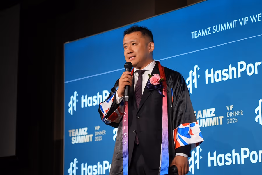 スーツと花飾りを着けた男性がマイクを持ち、『HashPort』のロゴが背景に表示されたステージで話している。