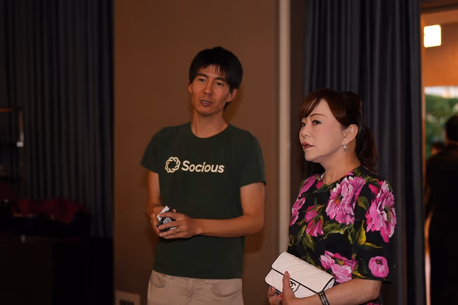 緑のTシャツに「Socious」と書かれた男性と、花柄のドレスを着て白いバッグを持つ女性が室内で話している。