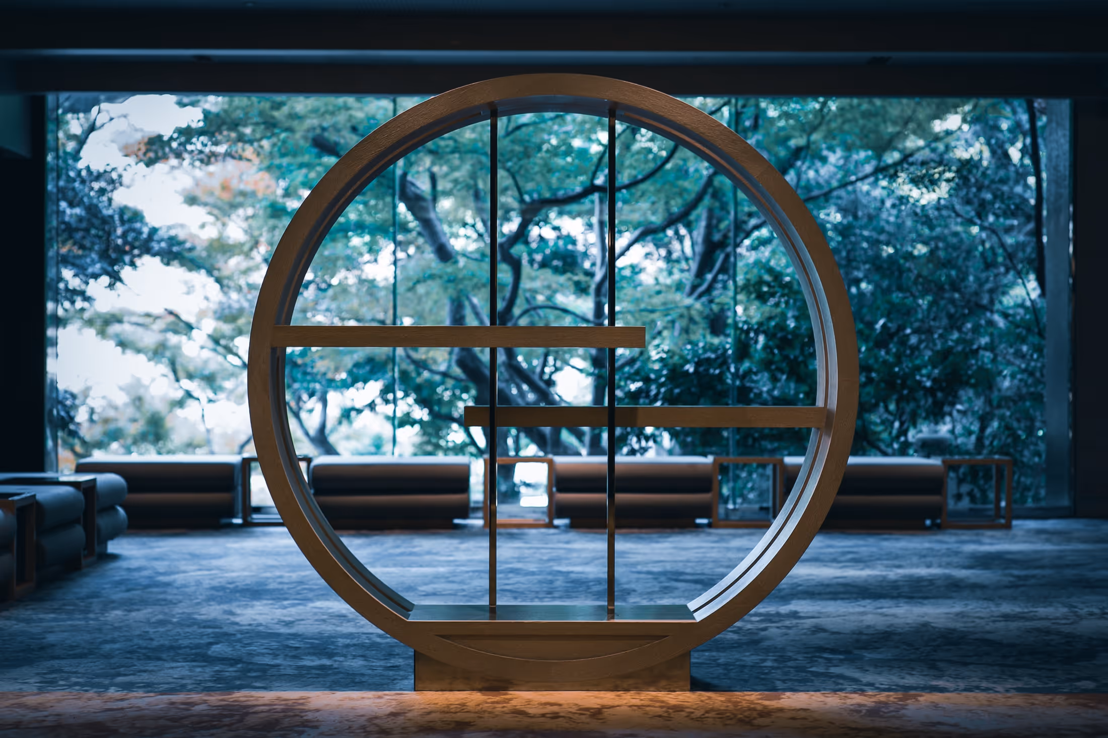 大型の丸い木製の飾り棚と、その背後に屋外の緑豊かな木々が見える室内空間。