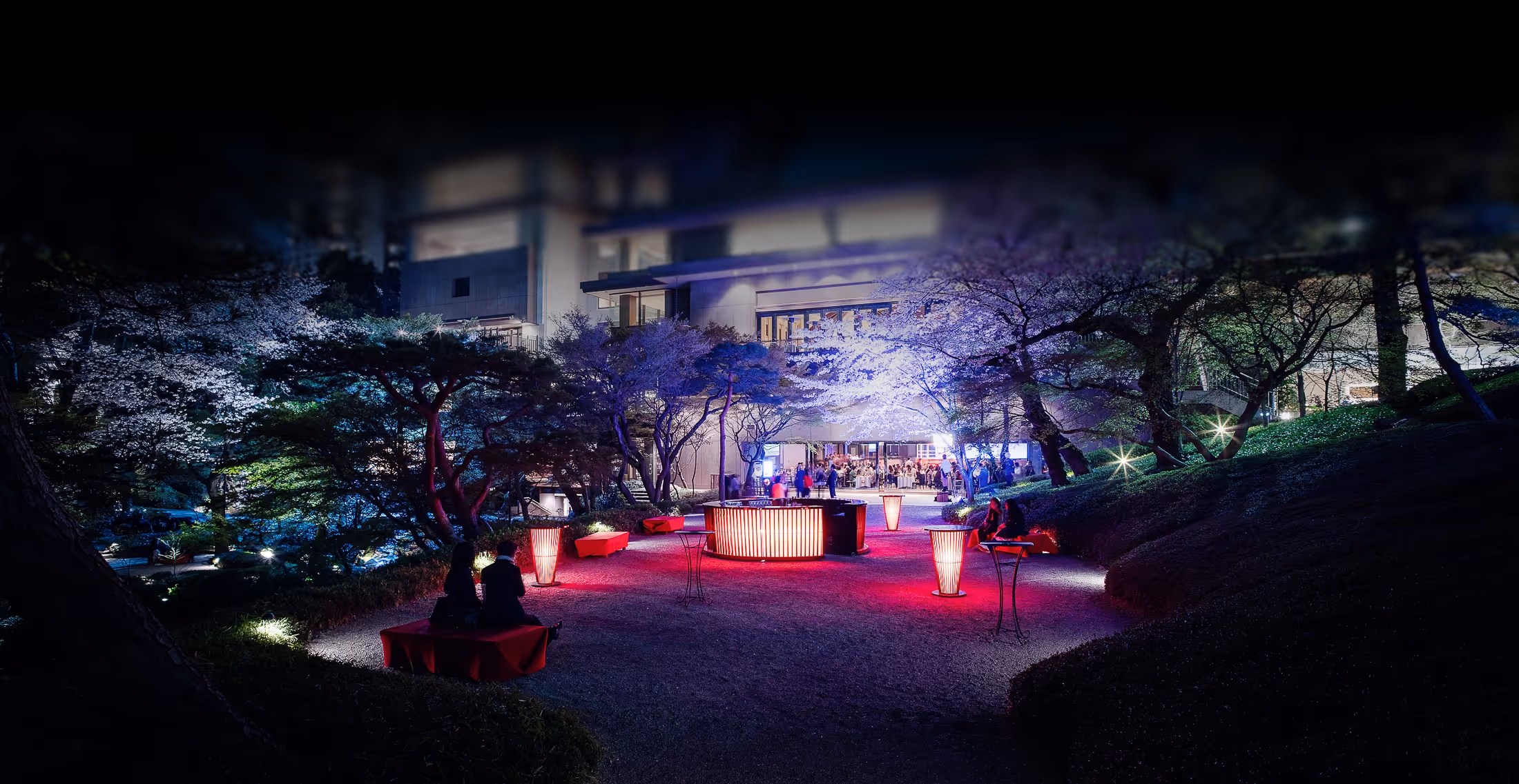 夜間にライトアップされた桜の木々に囲まれた庭園の中央に赤い光が灯るテーブルと椅子が設置され、多くの人が集まっている。