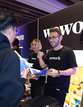 weworkのロゴが背景にあるブースで、黒いweworkのTシャツを着た男女が来場者に資料を配って説明している様子。