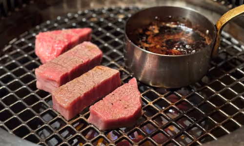 網の上で焼かれている厚切りの赤身肉のステーキと脂の入った小さな鍋。