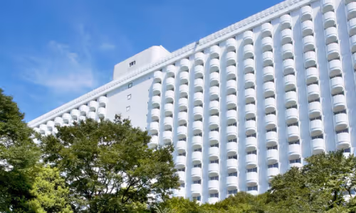 青空の下、緑の木々の前に立つ白いバルコニーが特徴の大型ホテル建物。