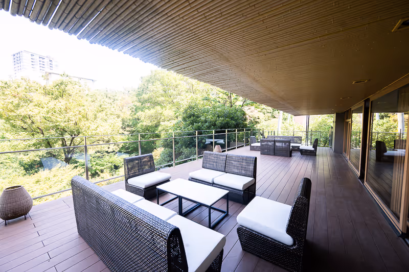 緑豊かな景色を望むウッドデッキに白いクッションと黒い編み込みの家具セットが並ぶ屋外ラウンジエリア。