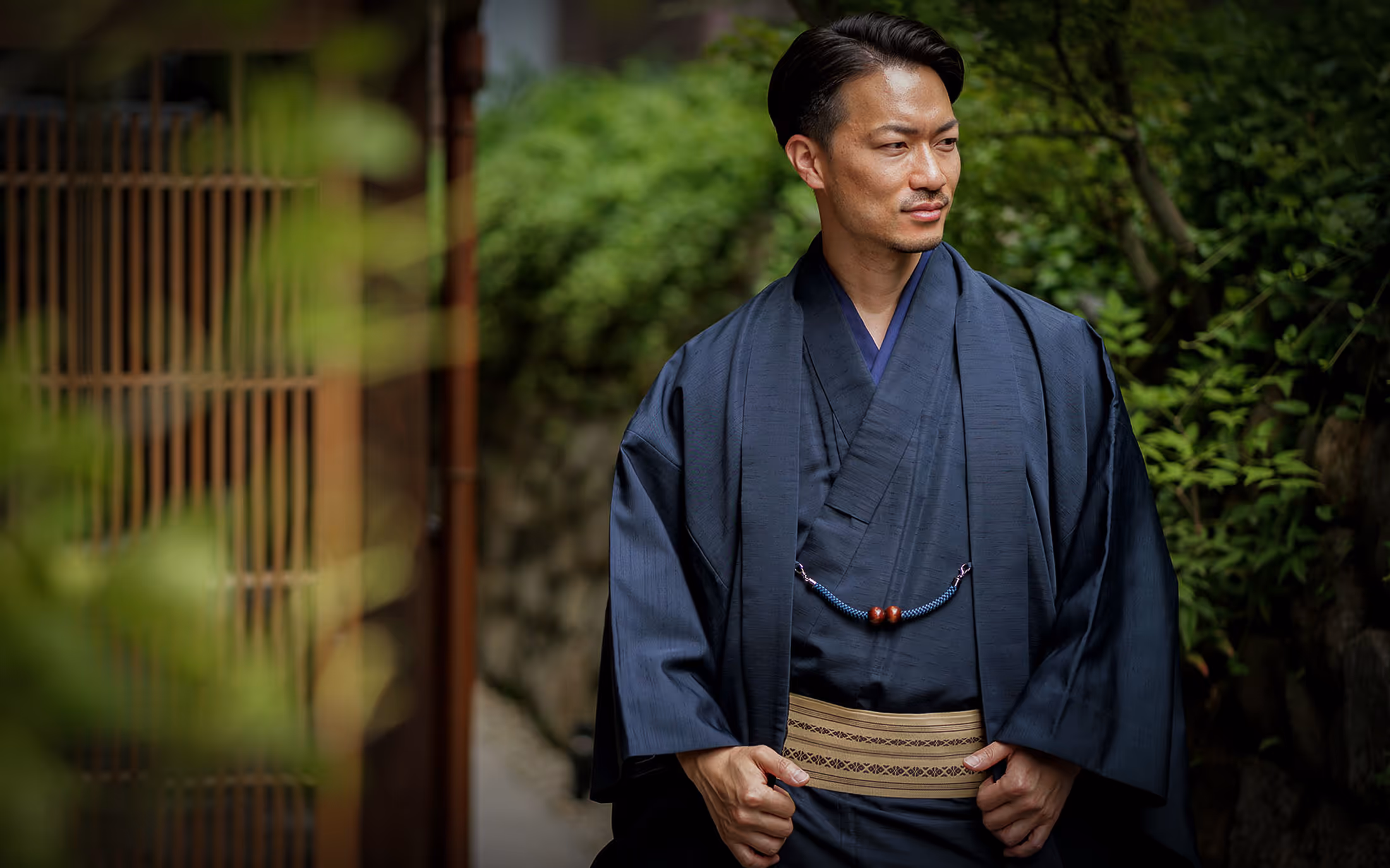 青い紋付き羽織袴を着て、庭園の通りを歩く日本人男性。