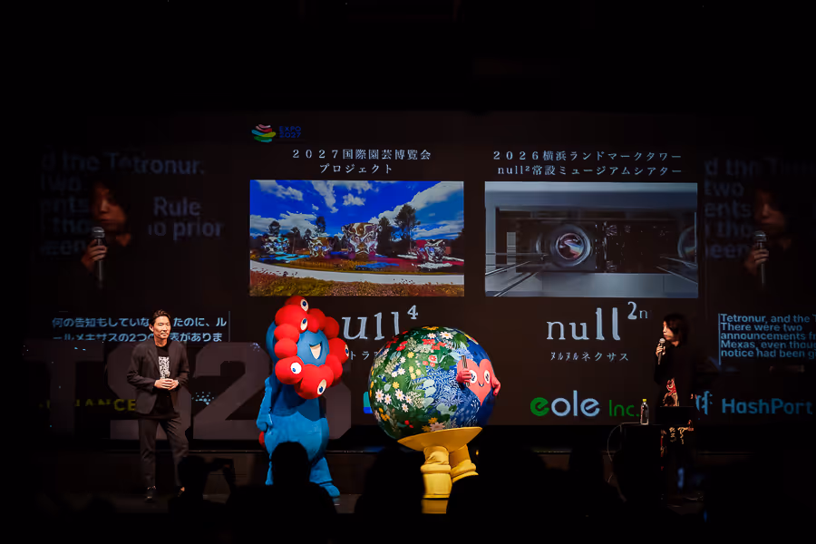 舞台でプレゼンテーション中の二人の人物と2体のカラフルなマスコットキャラクター、背景に2027年と2026年のプロジェクトを示すスクリーン。