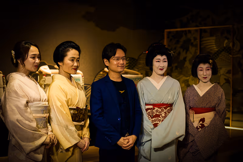 着物を着た4人の女性と紺色のジャケットを着た男性が一列に並び、背景は和風の装飾で照明が落ちている。