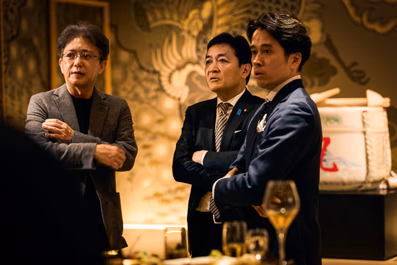 スーツとジャケットを着た3人の男性が腕を組みながら、室内の華やかな背景の前で真剣な表情で立っている。