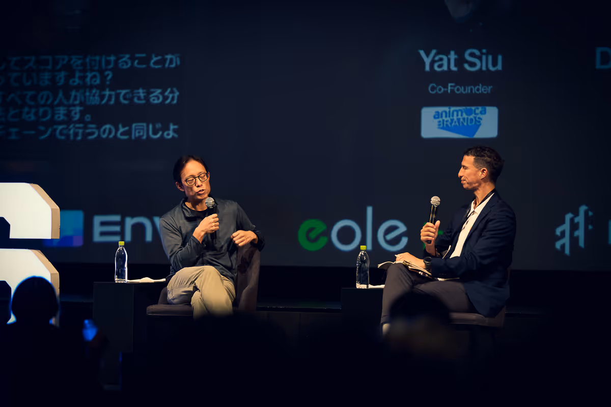 ステージでマイクを持って対話している二人の男性、背景に英語と日本語の文字や企業ロゴが表示されている。