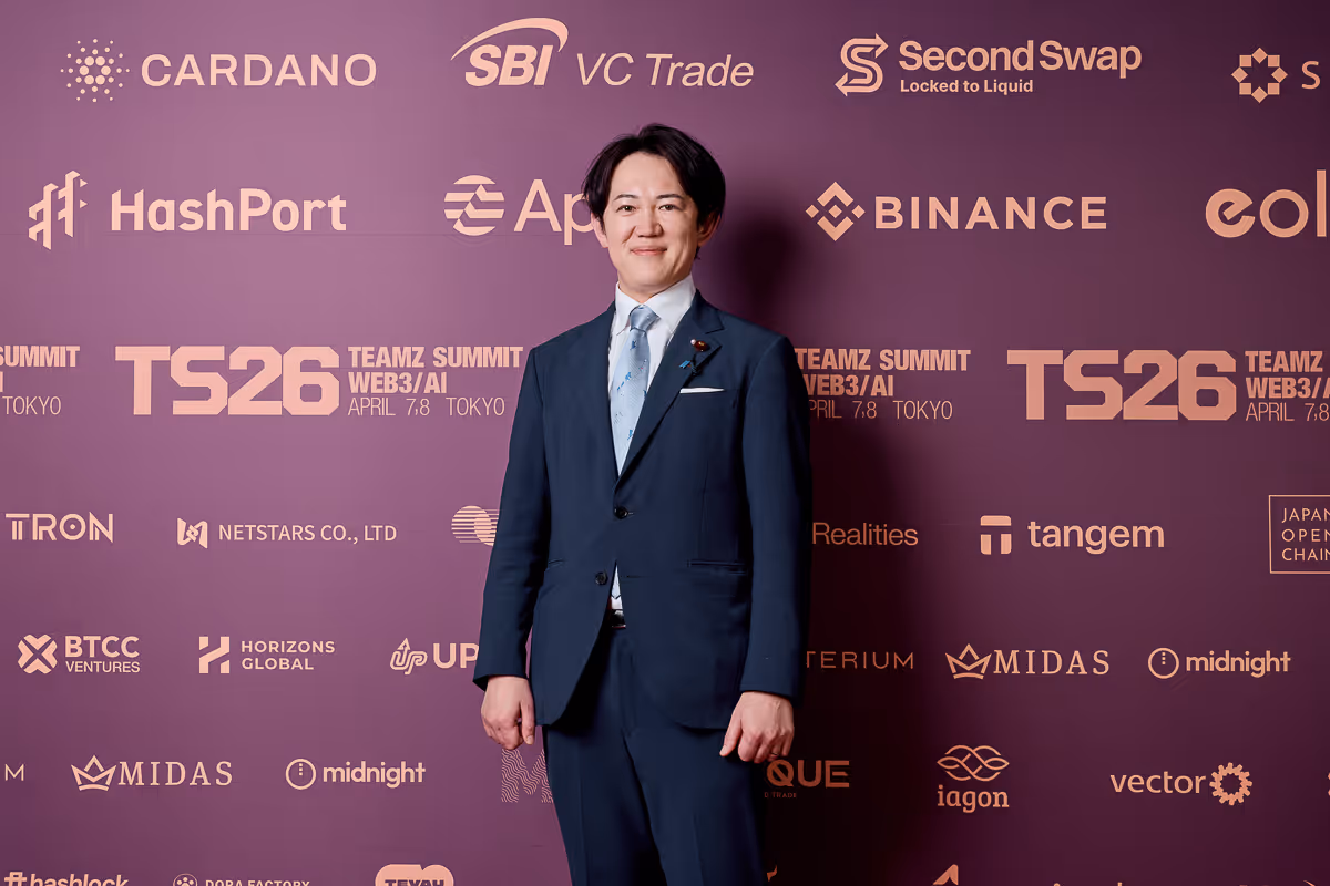 紺色のスーツを着た男性が、紫色の背景にさまざまなテクノロジー企業のロゴが並ぶ前で微笑んでいる。