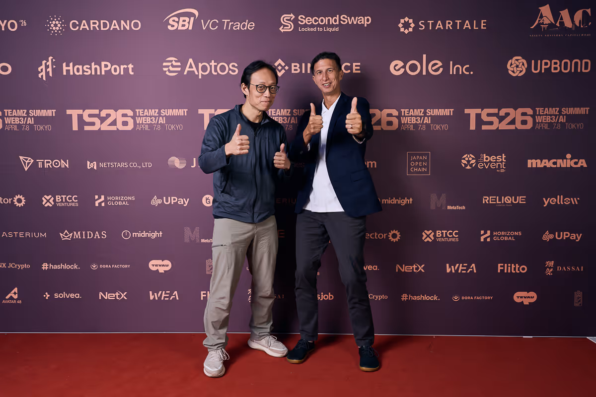 二人の男性が親指を立ててポーズをとり、背後に様々なテクノロジー企業のロゴが並ぶイベントの背景がある。