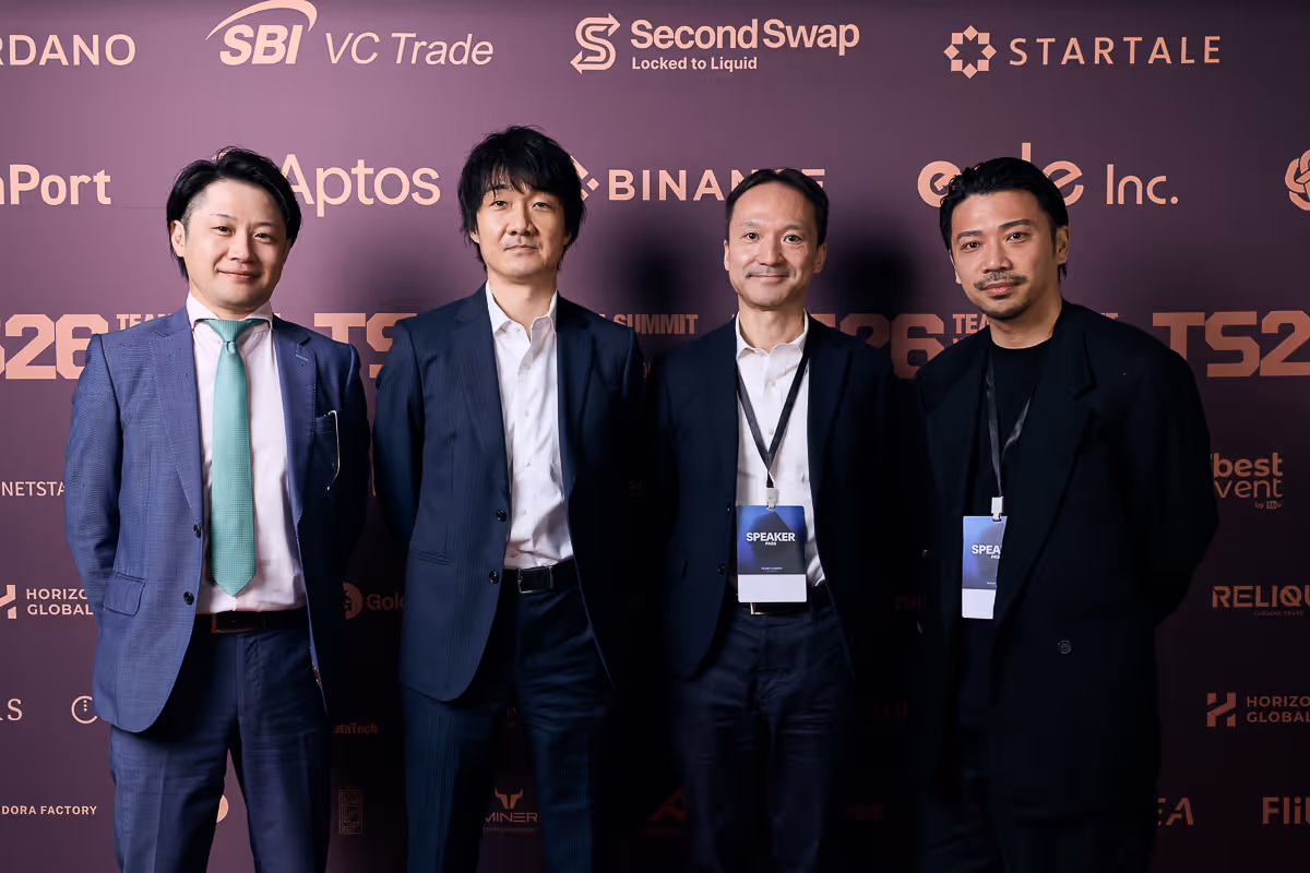 スーツを着た4人の男性が、仮想通貨関連のロゴが並ぶ背景の前で並んで立っている。