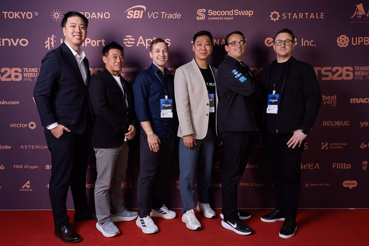 スーツやカジュアルな服装をした6人の男性が赤絨毯の前で並び、イベントバナーを背景に笑顔で立っている。