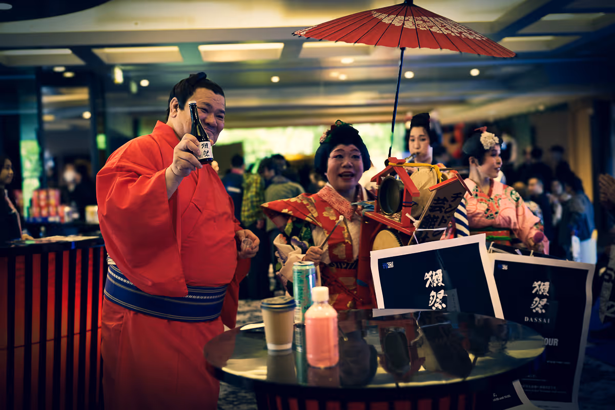 日本の伝統衣装を着た男性が日本酒のボトルを手に持ち、周囲に和服の女性たちがいる祝いの場面。