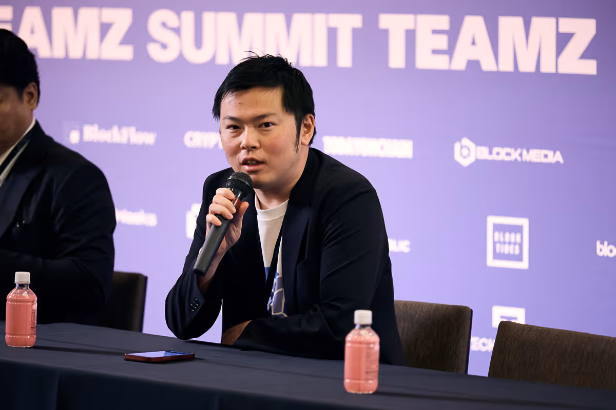 マイクを持って話しているビジネスカジュアルの男性が、紫色の背景にTEAMZ SUMMIT TEAMZの文字がある会議テーブルに座っている。