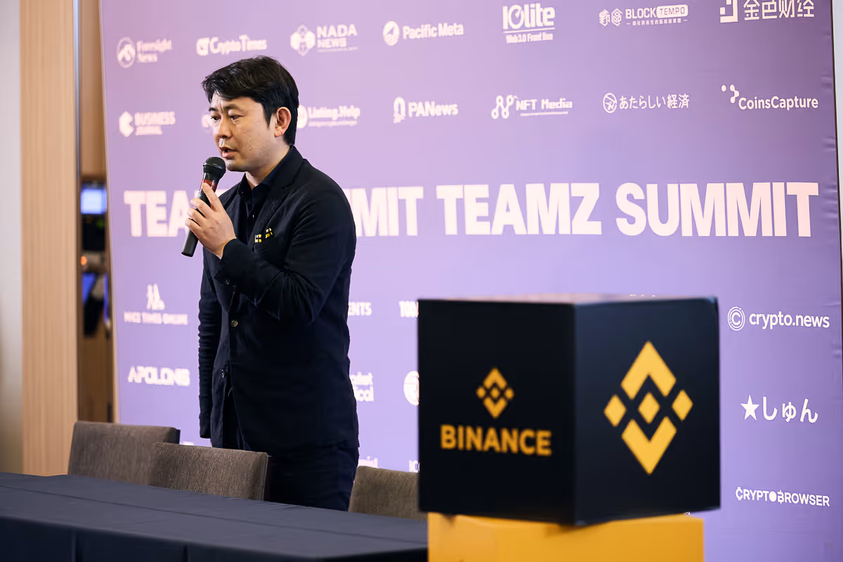 マイクを持ち、『TEAMZ SUMMIT』の背景の前で話すスーツ姿の男性。