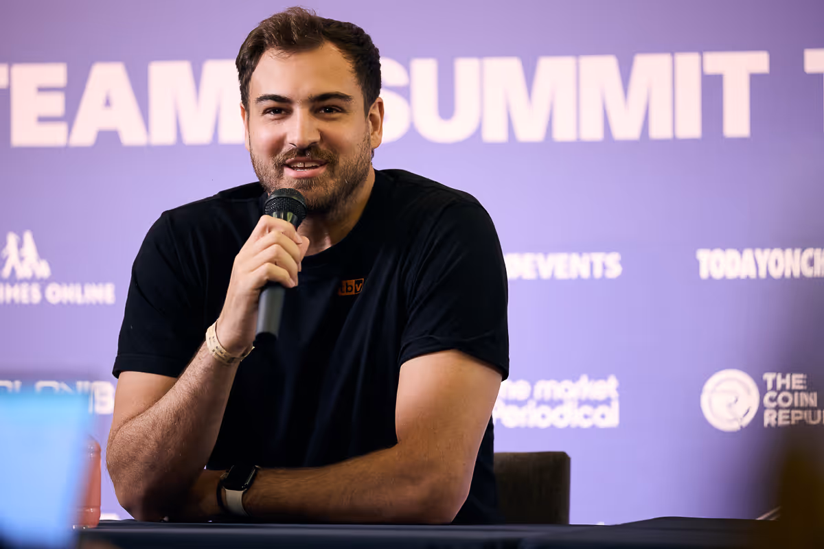 マイクを持って話している黒いTシャツの男性、背後に紫の背景とイベント名「TEAM SUMMIT」が見える。
