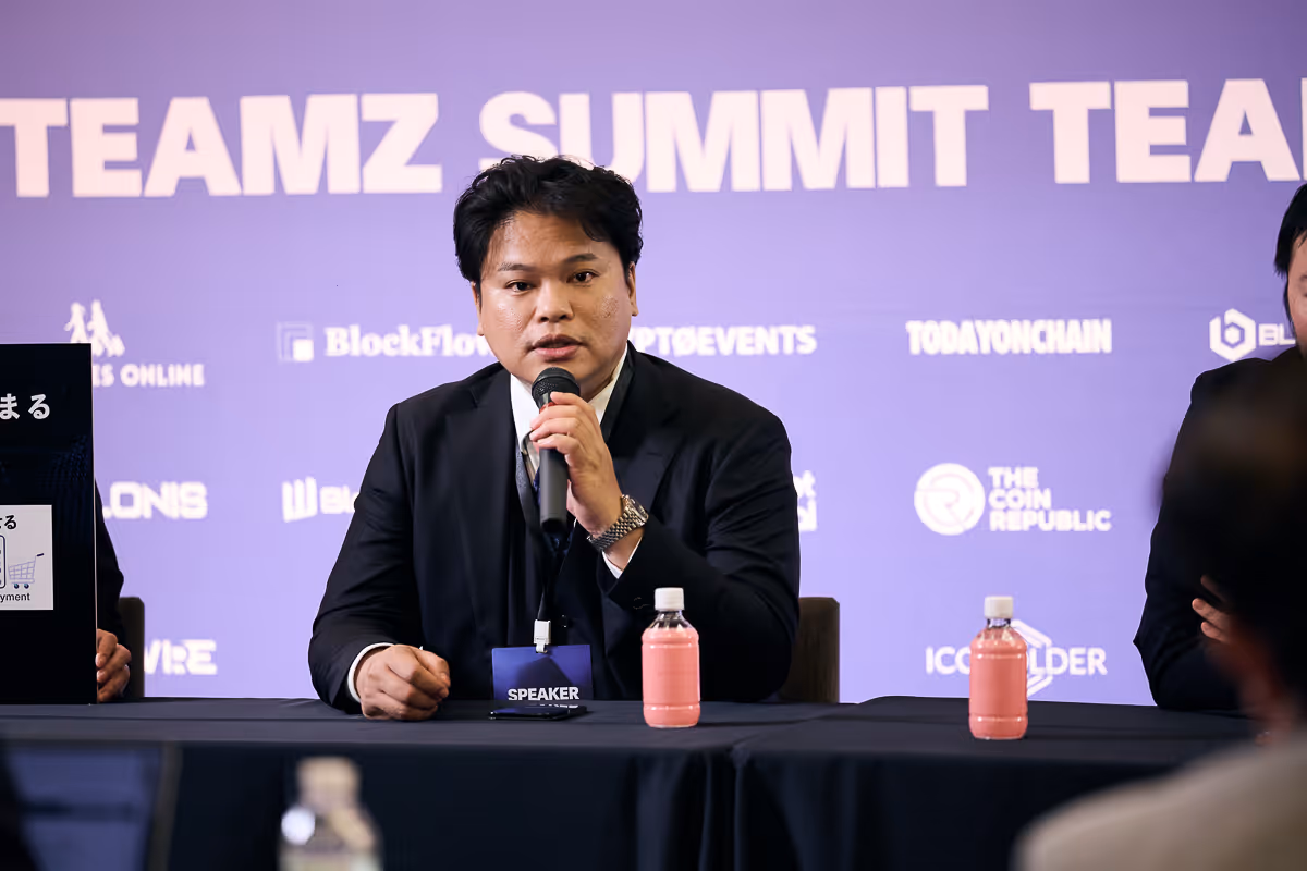 マイクを持ってスーツを着た男性が会議テーブルに座って話している。