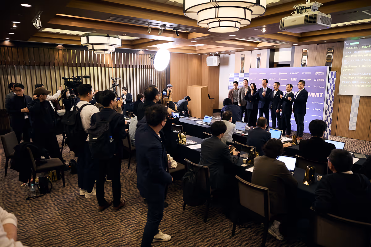 ビジネス会議でステージに並ぶ男女と、多くの参加者がノートパソコンを使い撮影や記録をしている様子。