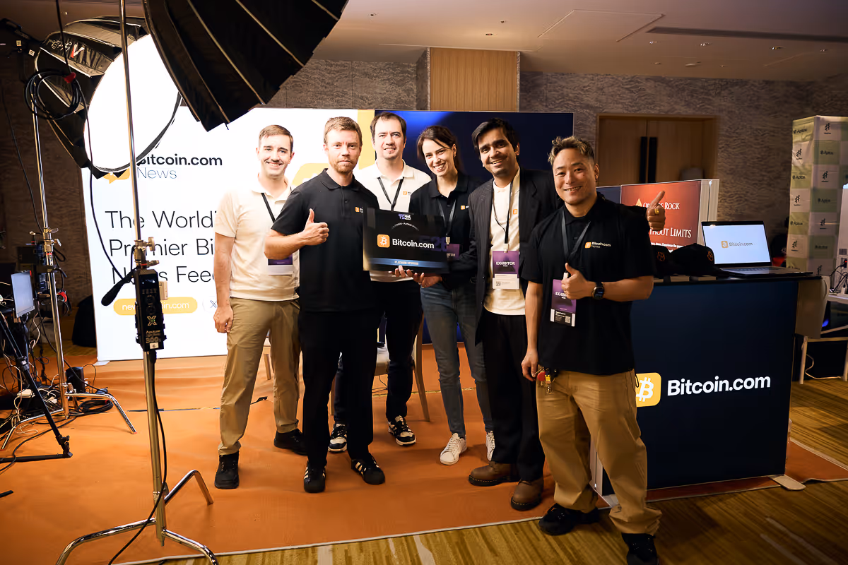 Bitcoin.comのブースでポーズを取る6人の男性、背景にBitcoin.comのロゴと展示パネル。