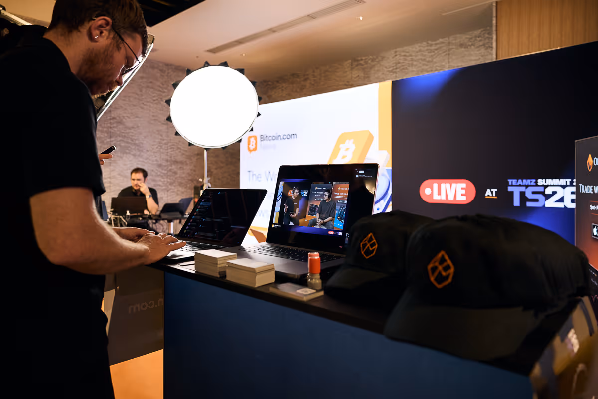 ノートパソコンの前で作業する男性とライブ配信中のビットコイン関連イベントのブース。