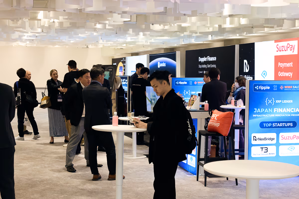 展示会でビジネス関連のディスプレイやブースを背景に、スマートフォンを操作する男性など複数の参加者がいる室内の様子。