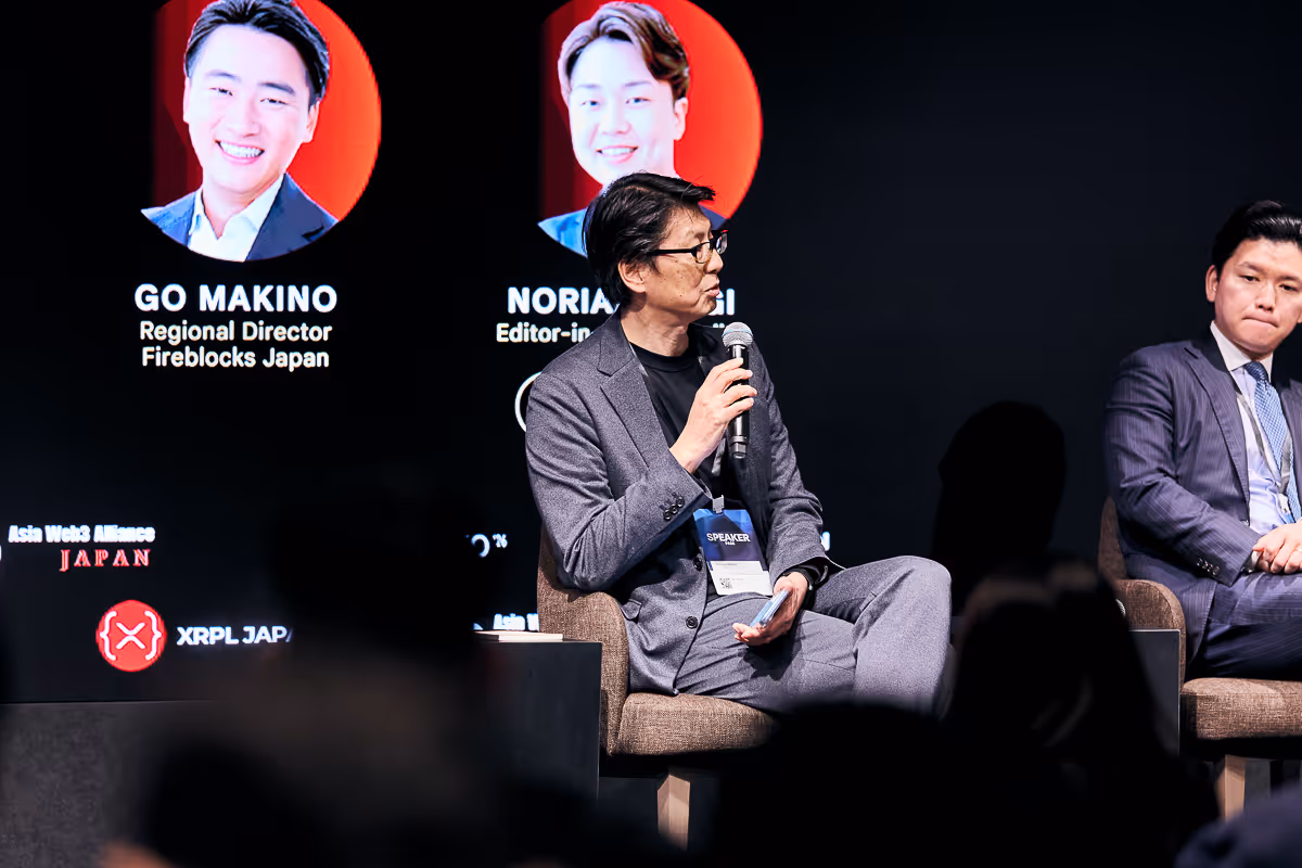 マイクを持ち座って話しているスピーカーと横に座る別の男性、背景にGo MakinoとNoria Sakaiの写真と肩書きが表示されたイベントのステージ。