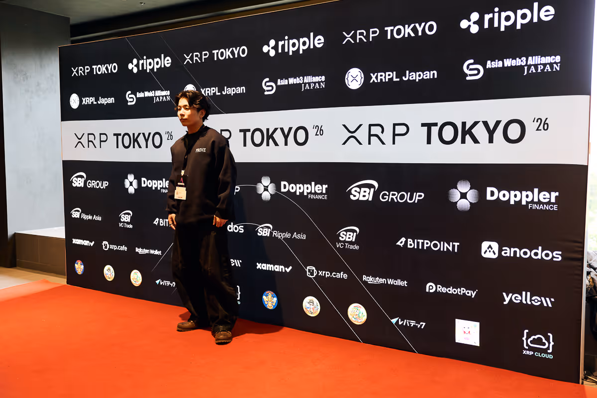 黒いパーカーを着た男性が赤いカーペットの上で、RippleやXRP TOKYO、SBIグループなどのロゴが並ぶ壁の前に立っている。