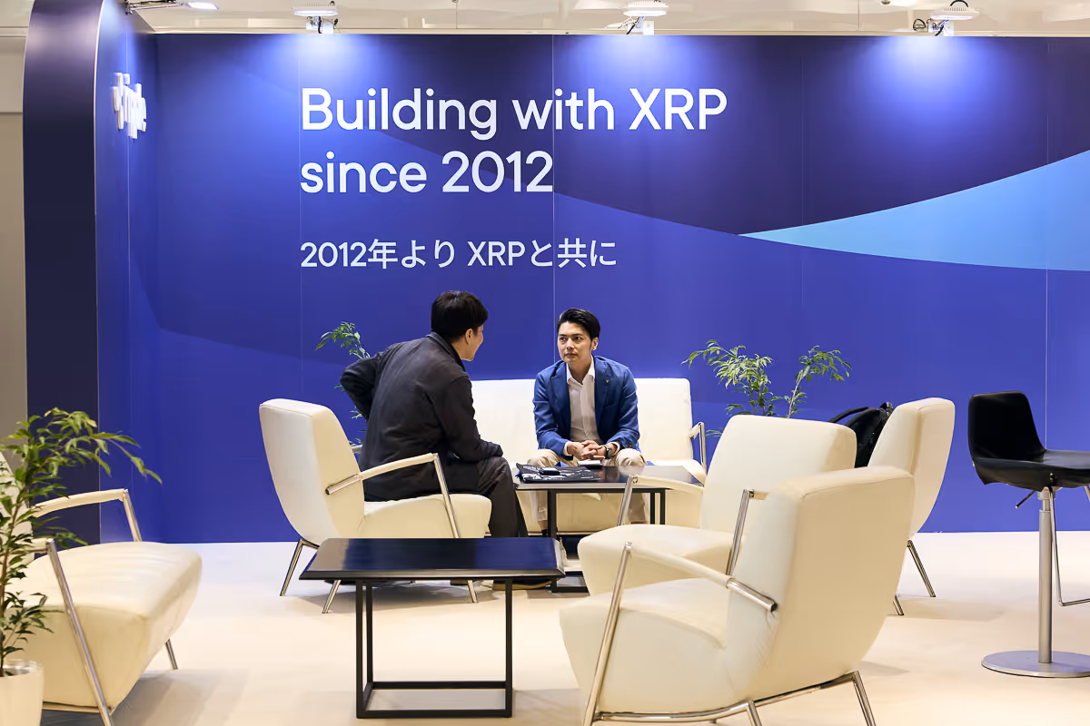 青い背景に「Building with XRP since 2012」と書かれた壁の前で、白いソファに座り向き合って話す二人の男性。
