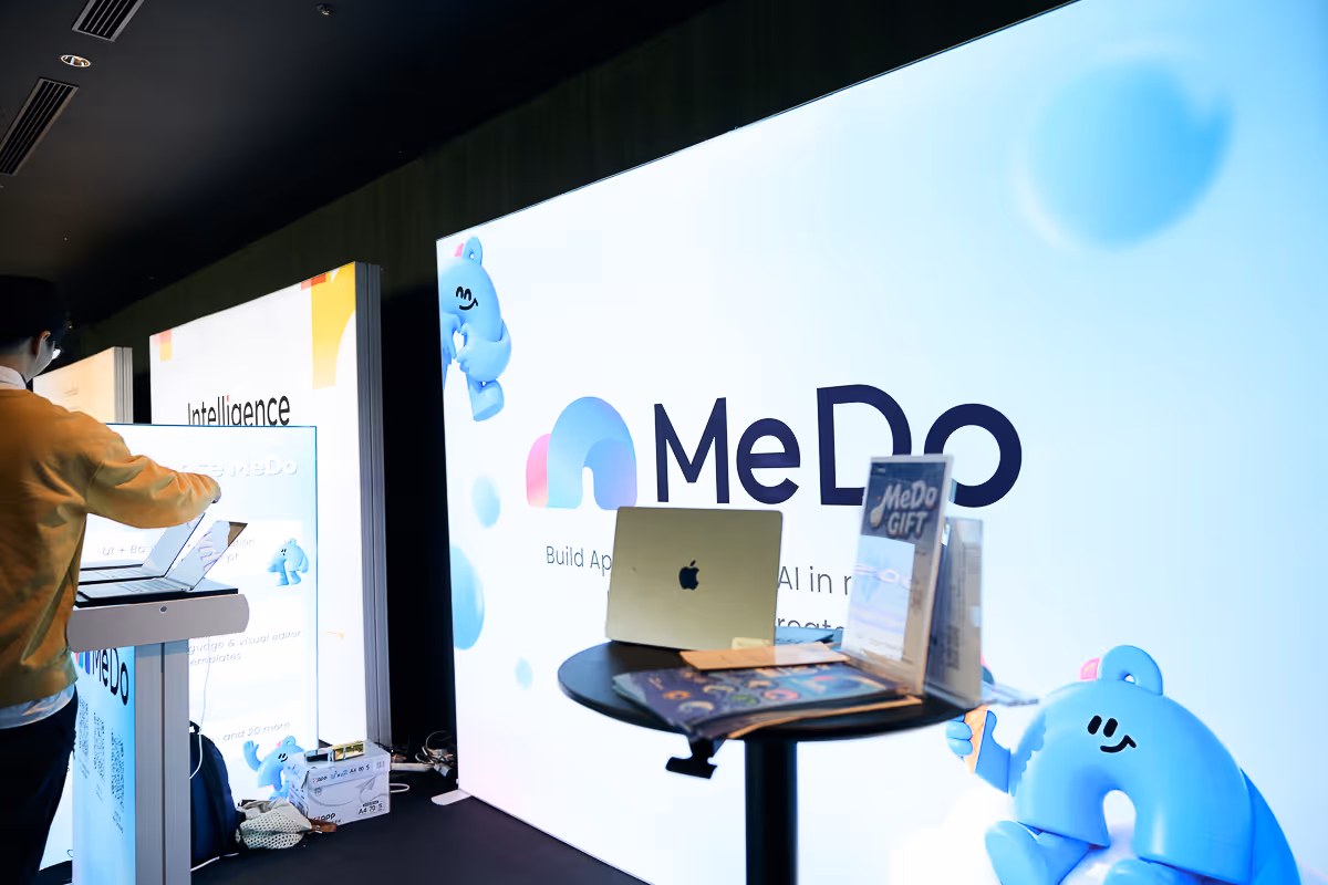 MeDOのロゴが表示された大型ディスプレイの前に、ノートパソコンとプロモーション資料が置かれたテーブルがあり、左側で人が操作している展示ブースの様子。