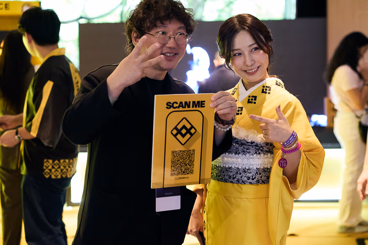 男性と着物を着た女性がQRコード付きの黄色いサインを持ち、笑顔でカメラに向かって指をさしている。