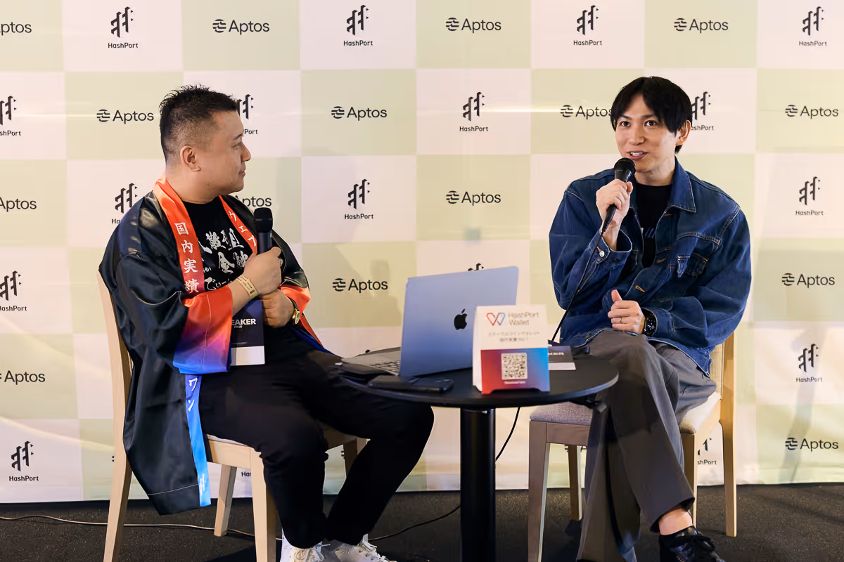 2人の男性がマイクを持ち、HashPortとAptosのロゴが背景にあるテーブルを挟んで対話している。