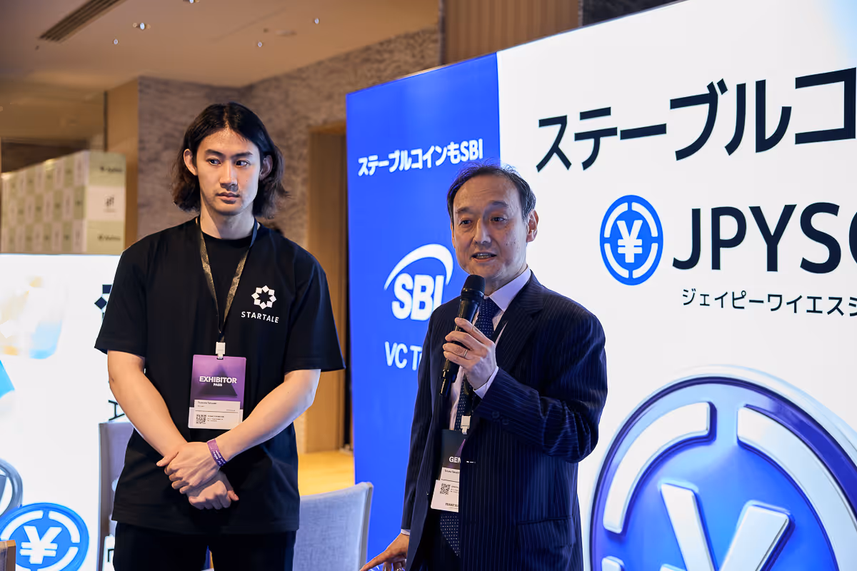 マイクを持ちスーツ姿で話す男性と、展示パスをつけたSTATALEの黒いTシャツの男性。