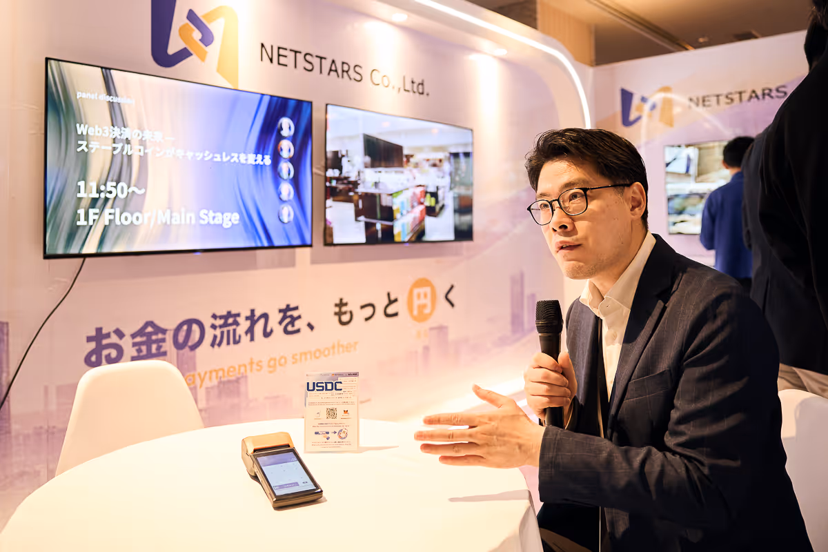 スーツを着た男性がマイクを持ち、NETSTARSの展示ブースで話をしている様子。