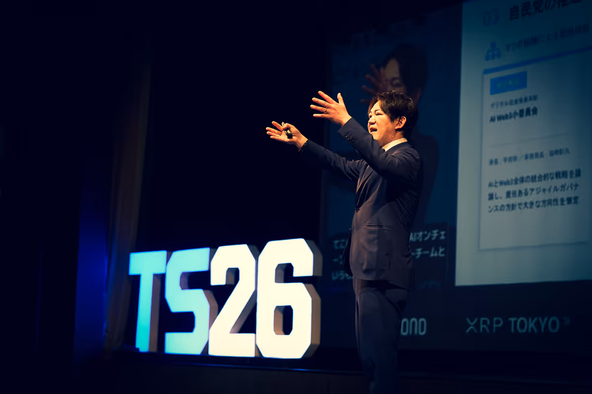 スーツを着た男性講演者が舞台でTS26の文字の横でプレゼンテーションをしている。