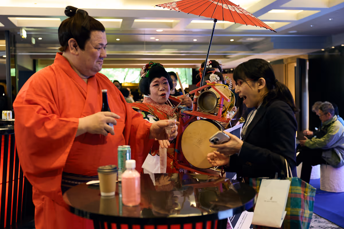 赤い着物を着た男性が女性に飲み物を差し出し、背景で楽器を持った人物と赤い和傘が見える。