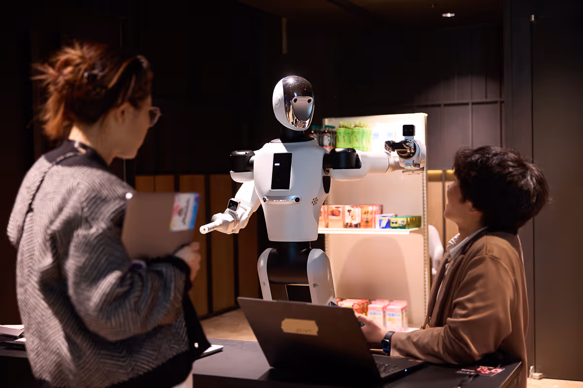 ラップトップを使用している男性とメモ帳を持つ女性の前で腕を広げる白黒ロボット。